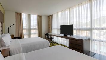 A hotel room with two beds, a desk with a television, and large windows with sheer curtains.
