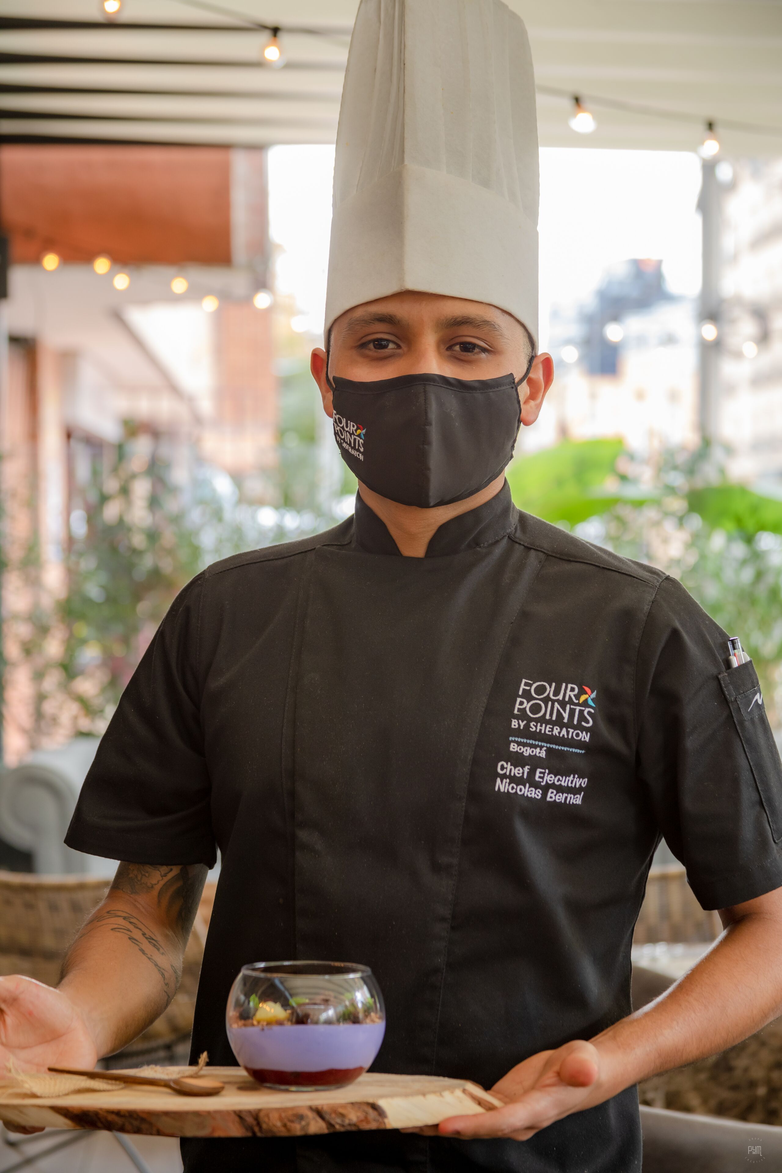 A chef in a black uniform with a white hat, holding a wooden board with a bowl of food.