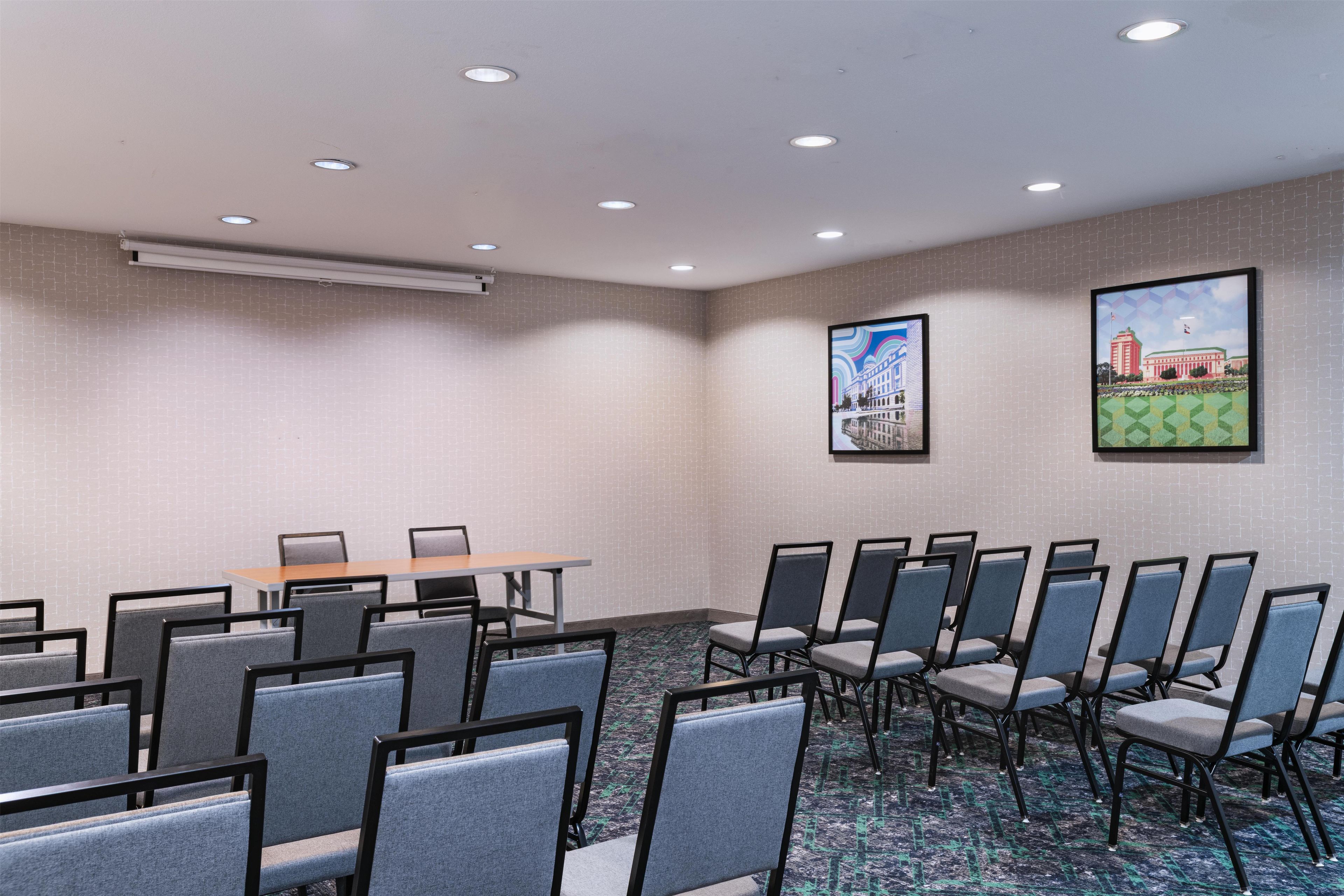 Un salón de conferencias con una larga mesa y filas de sillas, aire acondicionado y dos cuadros en la pared.