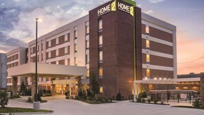 A multi-story hotel with a prominent "HOME 2 HOME" sign, featuring a modern design with brick and glass facade, a covered entrance, and a landscaped outdoor area.