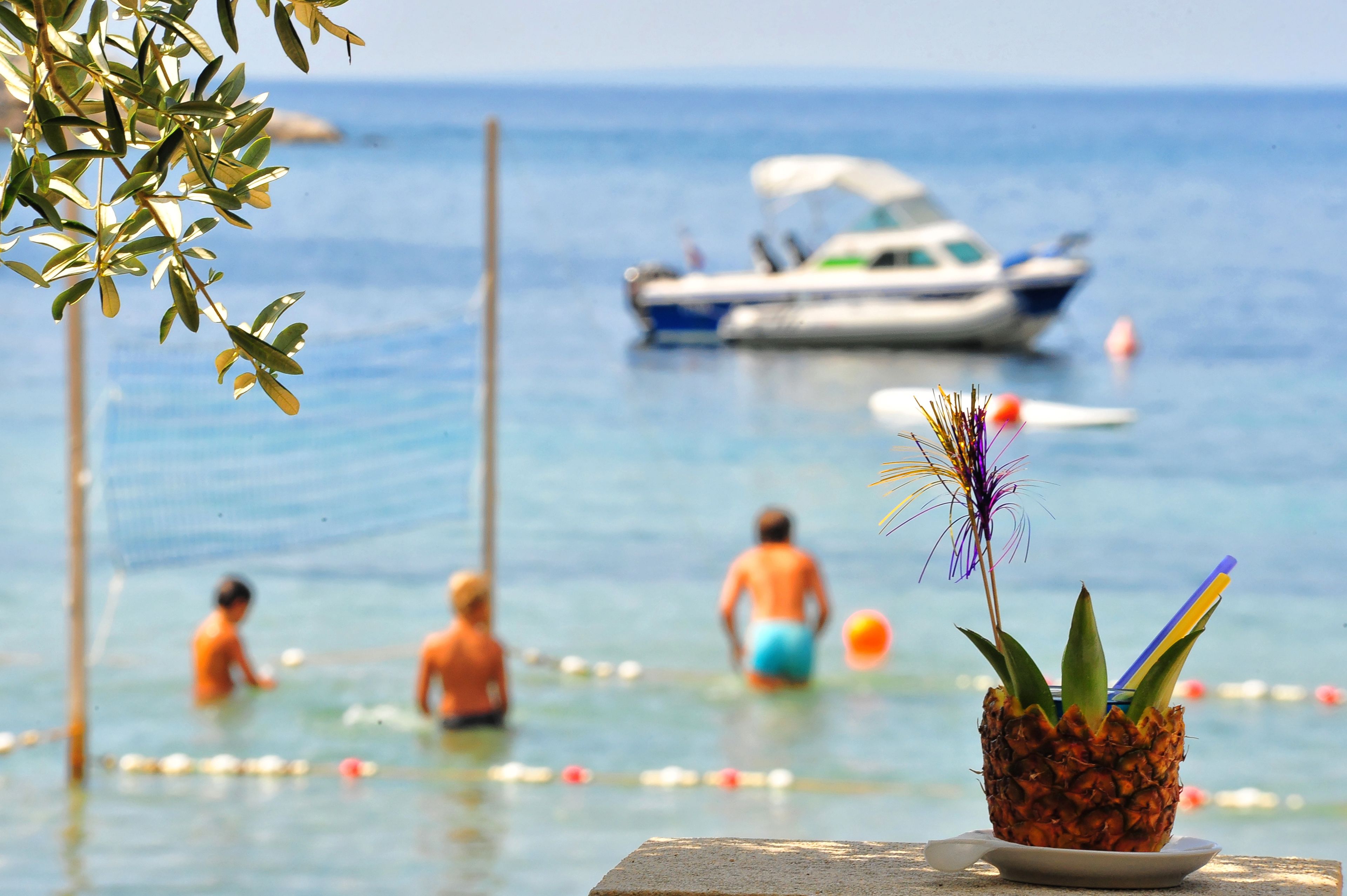 A beach scene with a pineapple on a table, people in the water, and a boat in the background.