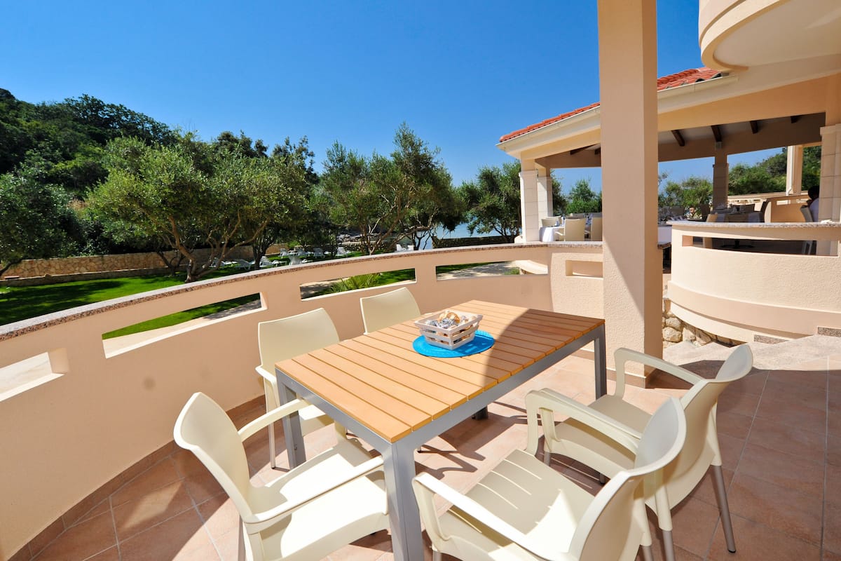 A covered outdoor patio with a table and chairs, overlooking a landscaped garden and a body of water.
