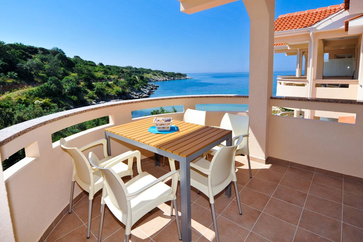 A balcony with a wooden table and white chairs, overlooking a coastal view with a clear sky.