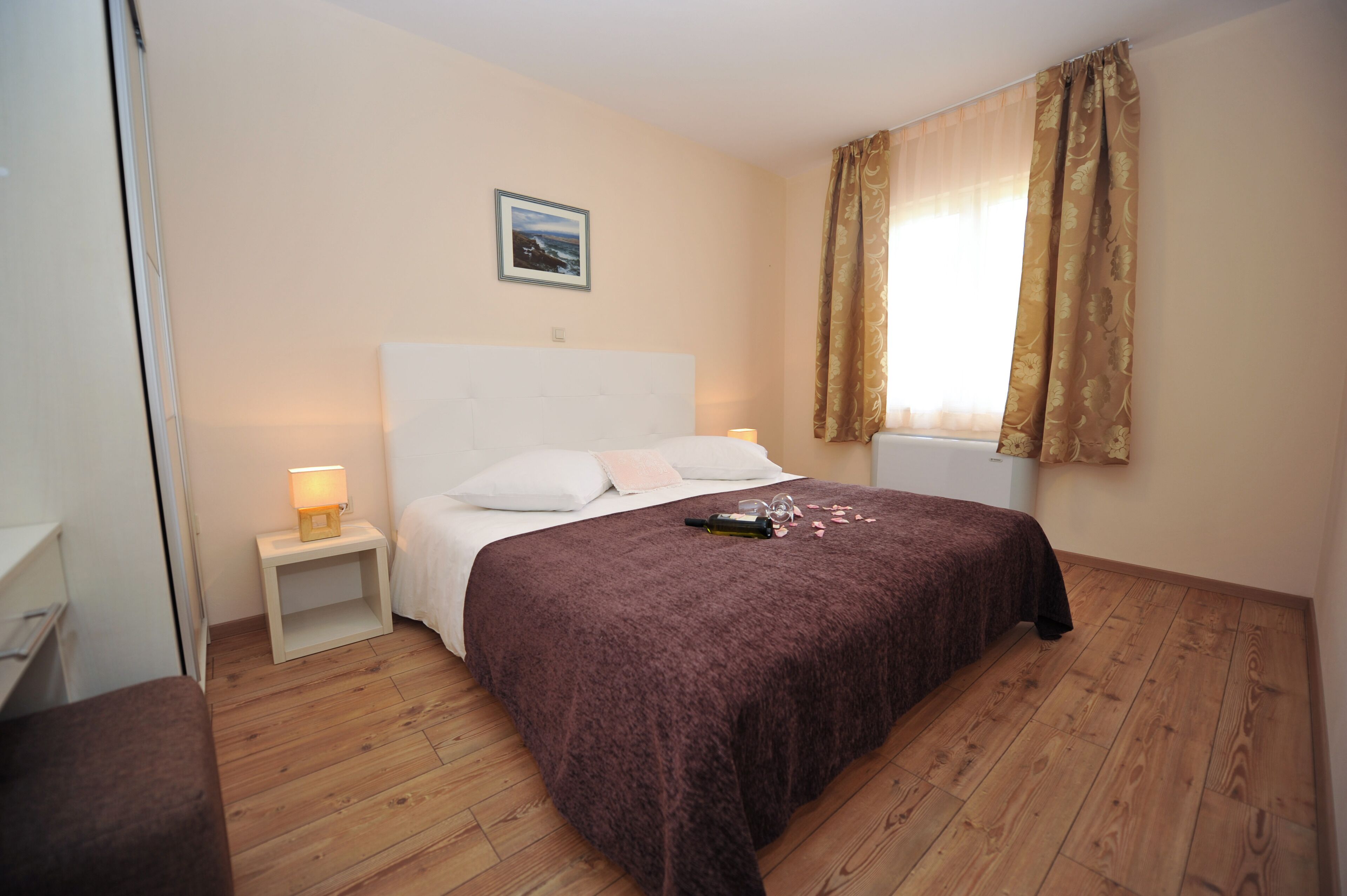 A bedroom with a wooden floor, a white headboard, a bed with a brown blanket, a bedside table with a lamp, and a window with curtains.