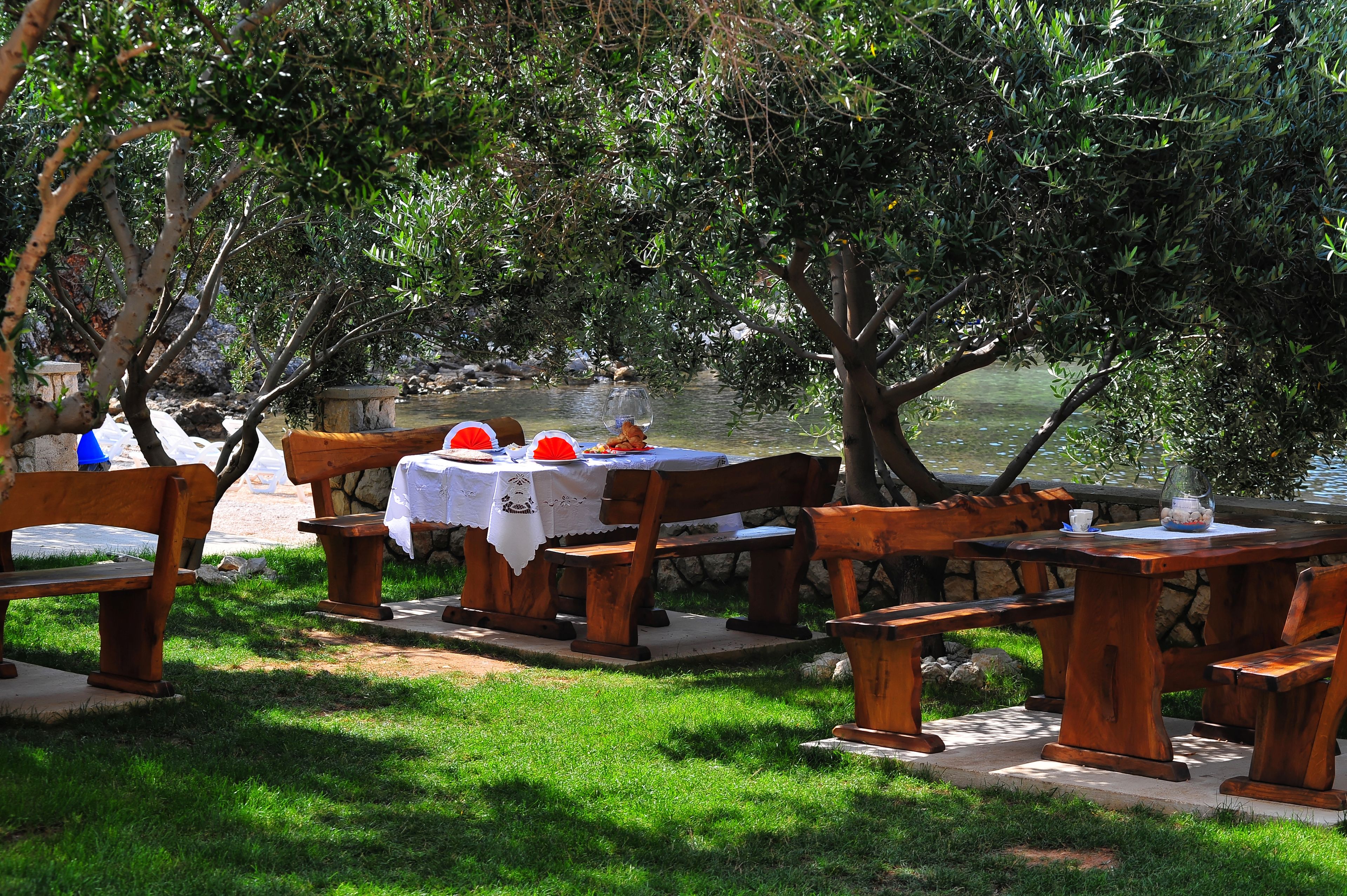An outdoor dining area with wooden benches and tables, surrounded by lush greenery and a body of water.