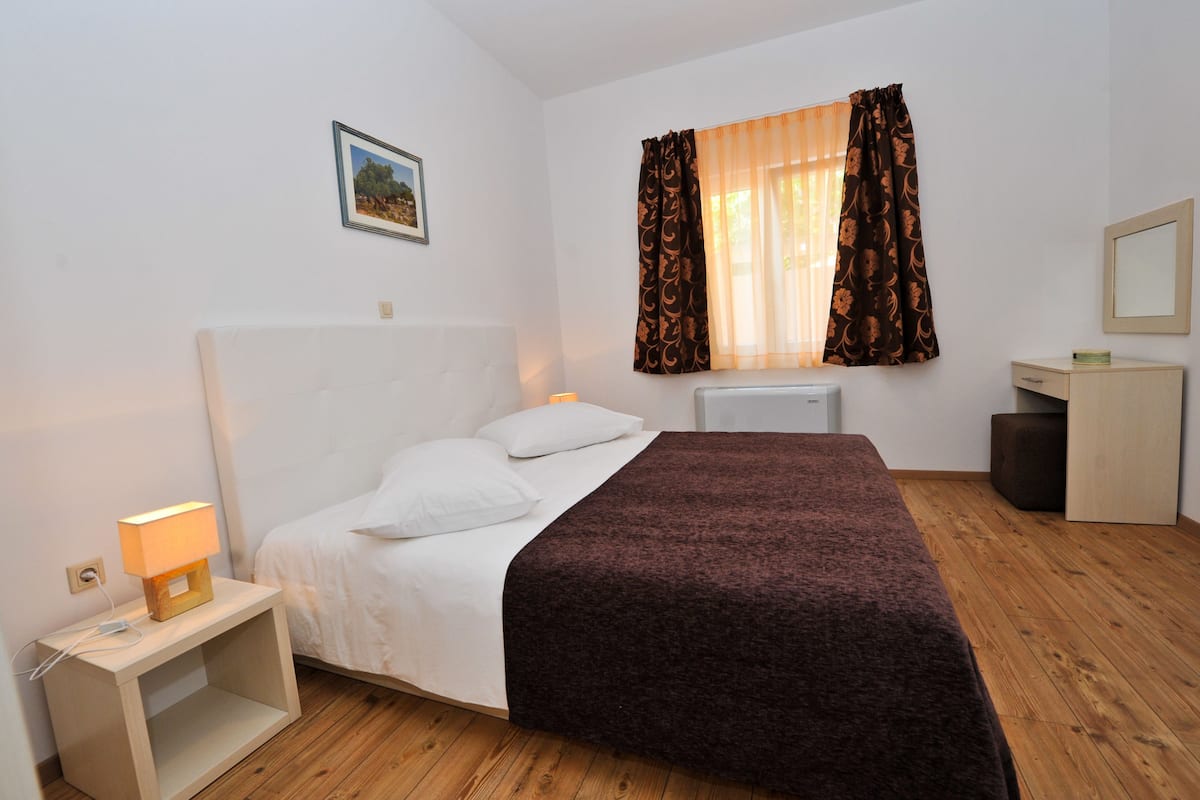 A bedroom with a bed, bedside table, lamp, window with curtains, and a small bedside cabinet.