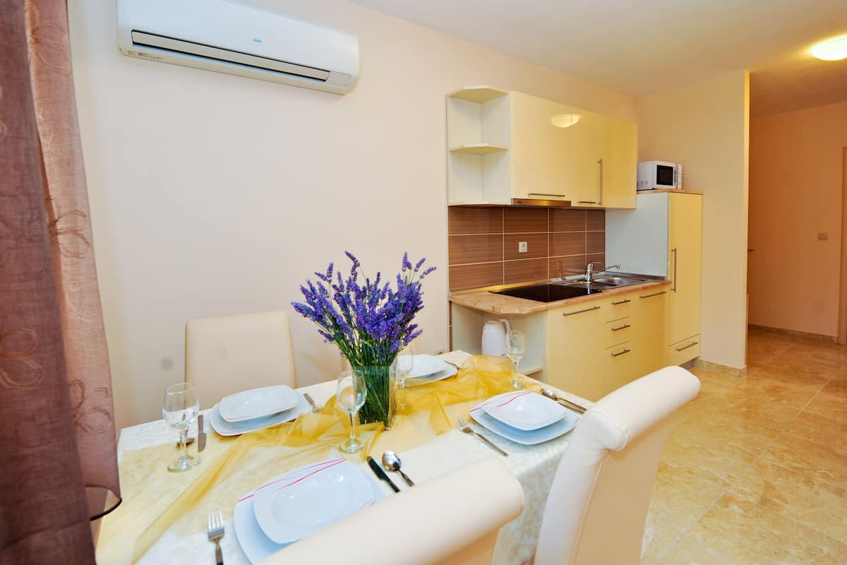 A dining area with a table set for four, a vase with purple flowers, and a kitchen in the background.