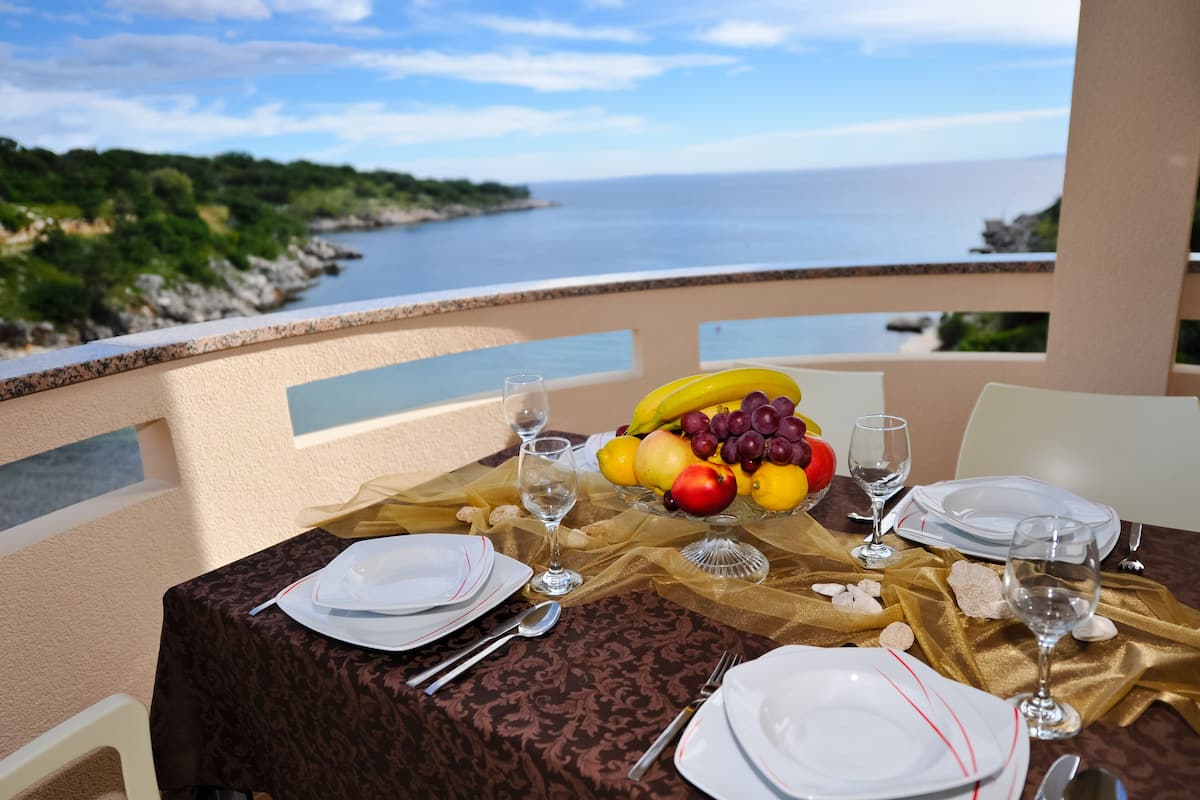 A table set for two with plates, cutlery, and a fruit bowl, overlooking a coastal view.