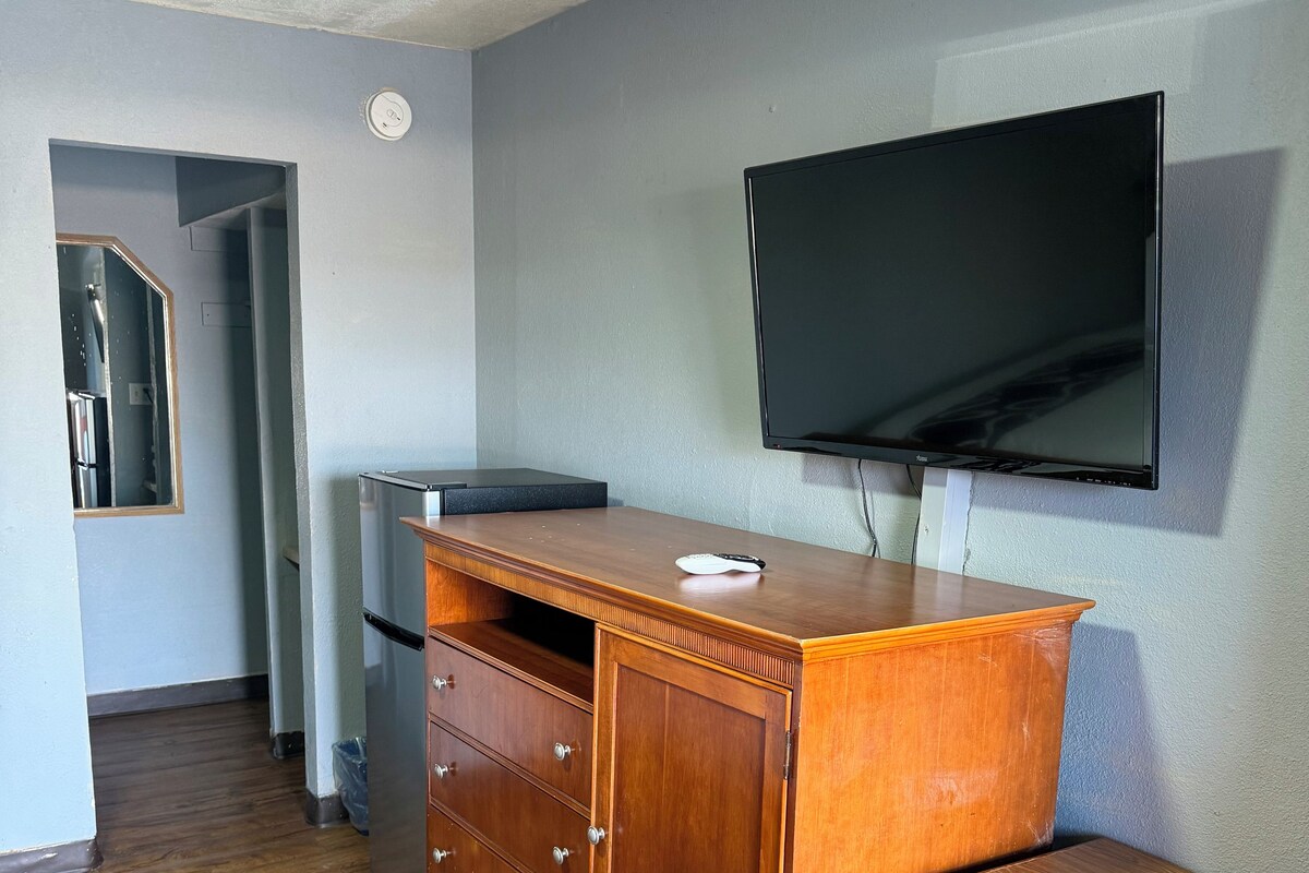 A room with a wooden dresser, a television mounted on the wall, a wooden table, and a chair with a cushion.