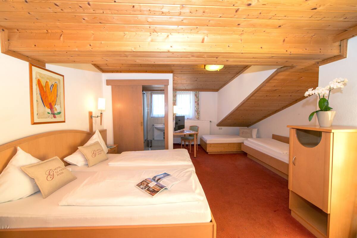 A hotel room with a wooden ceiling, a bed with white bedding, a bedside table with a lamp, a small desk with a chair, and a wooden cabinet.