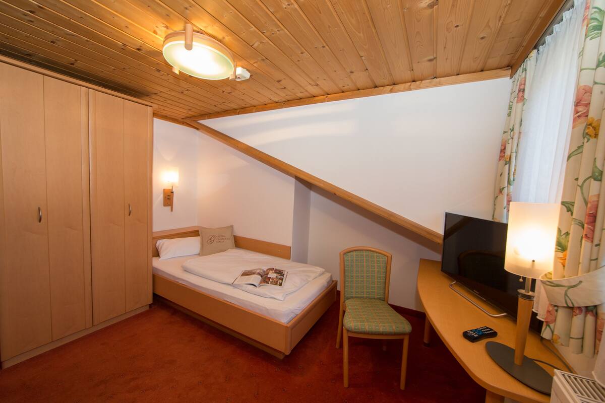 A bedroom with a wooden ceiling, a bed, a chair, a desk with a television, and a wardrobe.