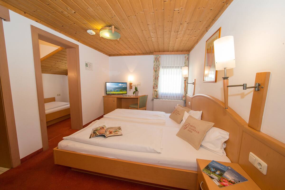 A hotel room with a wooden ceiling, a bed with white linens, a desk with a computer, and a window with curtains.