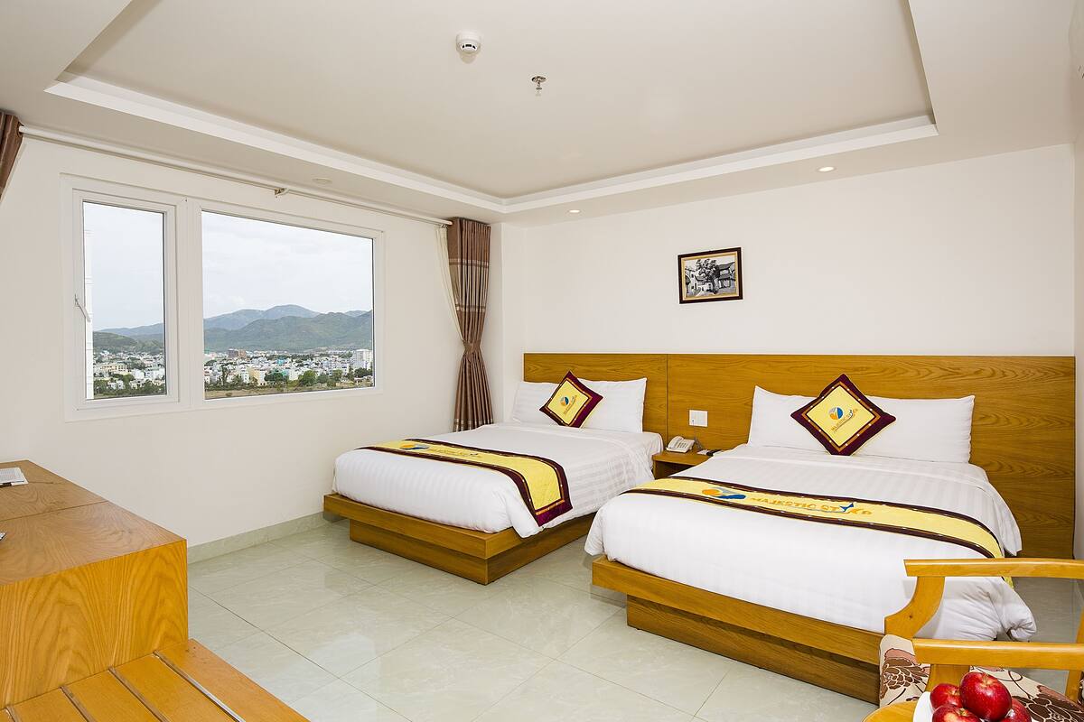 A hotel room with two beds, a desk, and a view of a city and mountains.