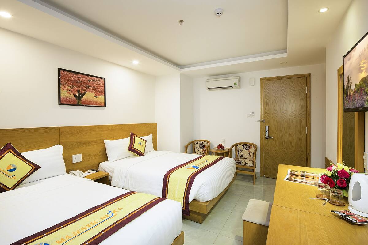 A hotel room with two beds, a wooden headboard, a desk with a chair, a small table with a vase of flowers, and a painting on the wall.