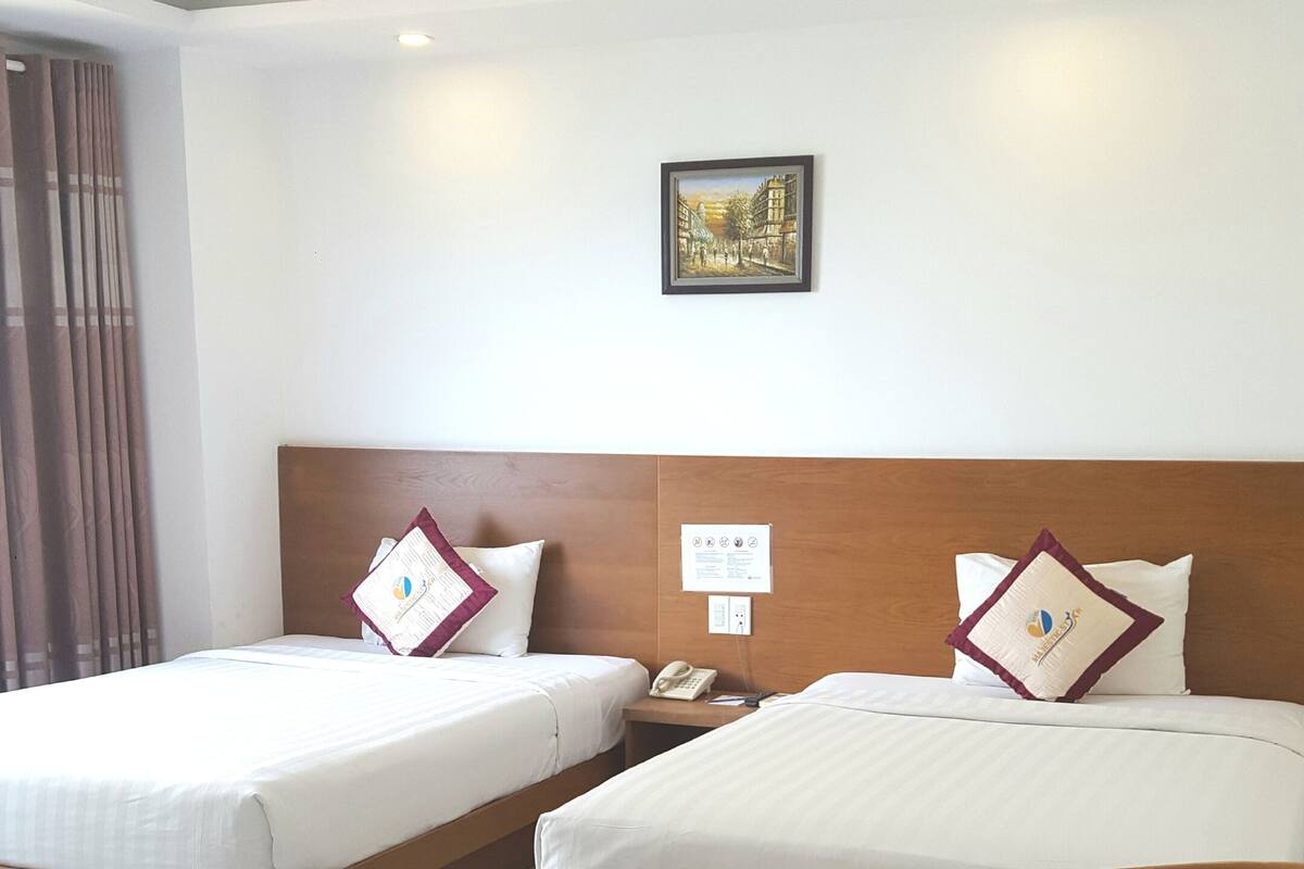 A hotel room with two beds, a wooden headboard, a bedside table with a phone, a framed picture on the wall, and a window with curtains.