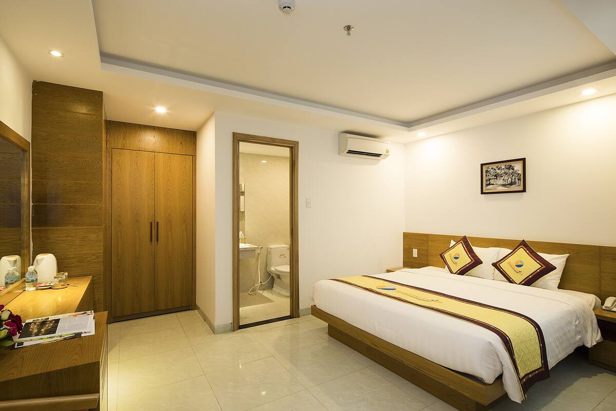 A hotel room with a wooden wardrobe, a bedside table with a lamp, a bed with a white comforter, and a framed picture on the wall.