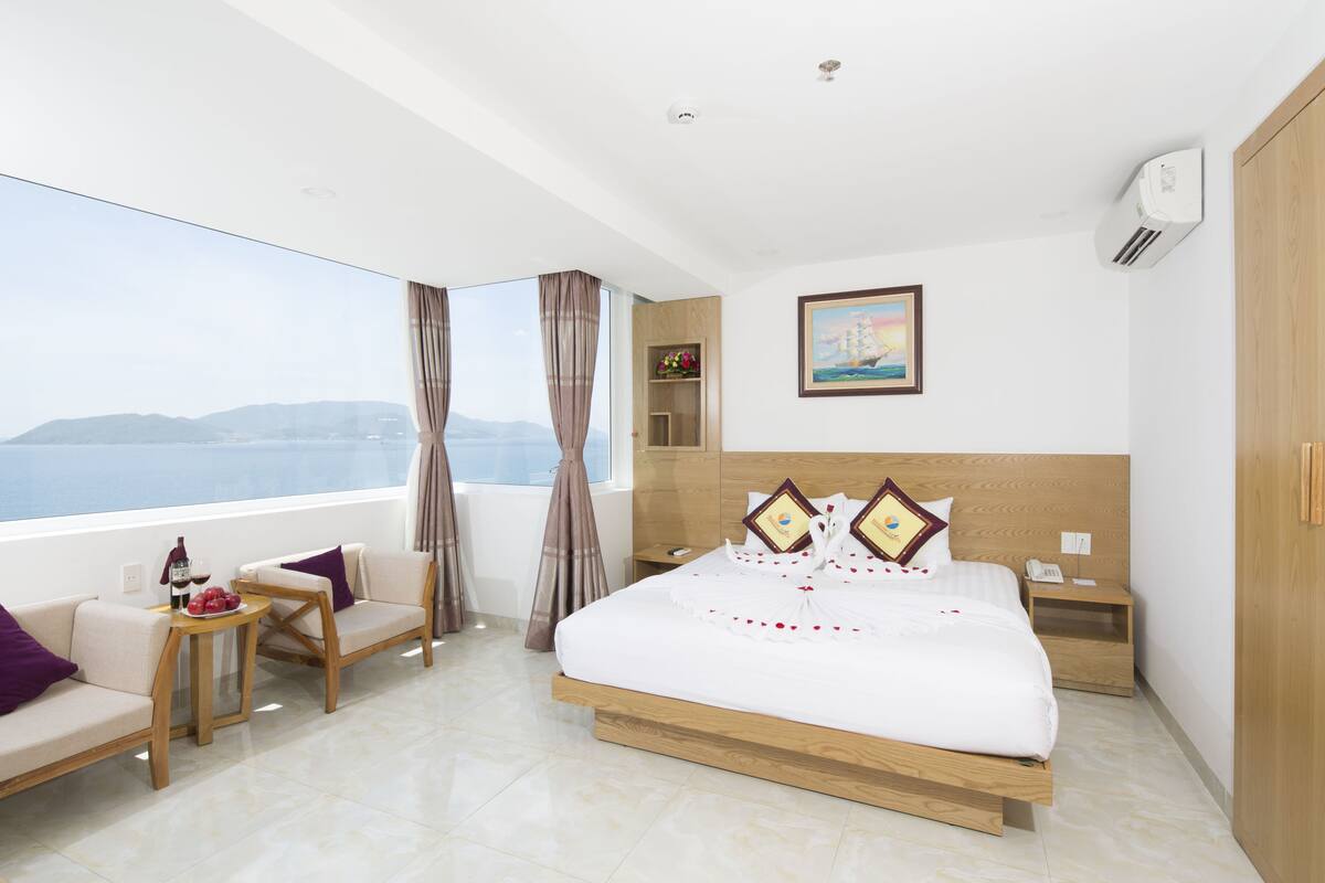 A modern hotel room with a large bed, a sofa, a small table, and a view of the sea.