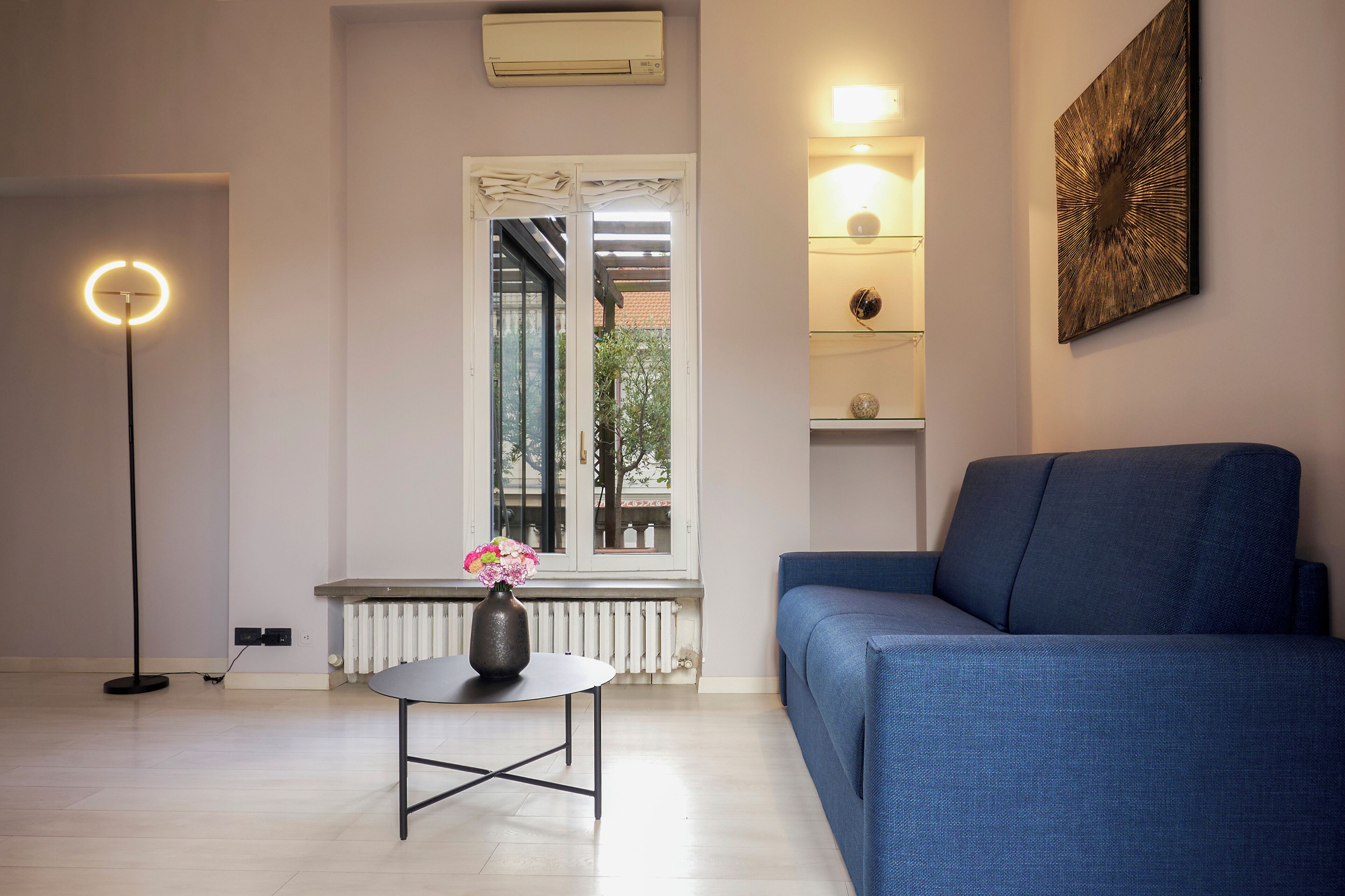 Una sala de estar moderna con un sofá azul, una mesa redonda con un florero, una lámpara de pie y un radiador.