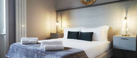 Un lit bien fait, recouvert d’une couvre-lit à motifs, deux tables de chevet avec des lampes, un miroir rond au-dessus du lit et une fenêtre avec des rideaux.