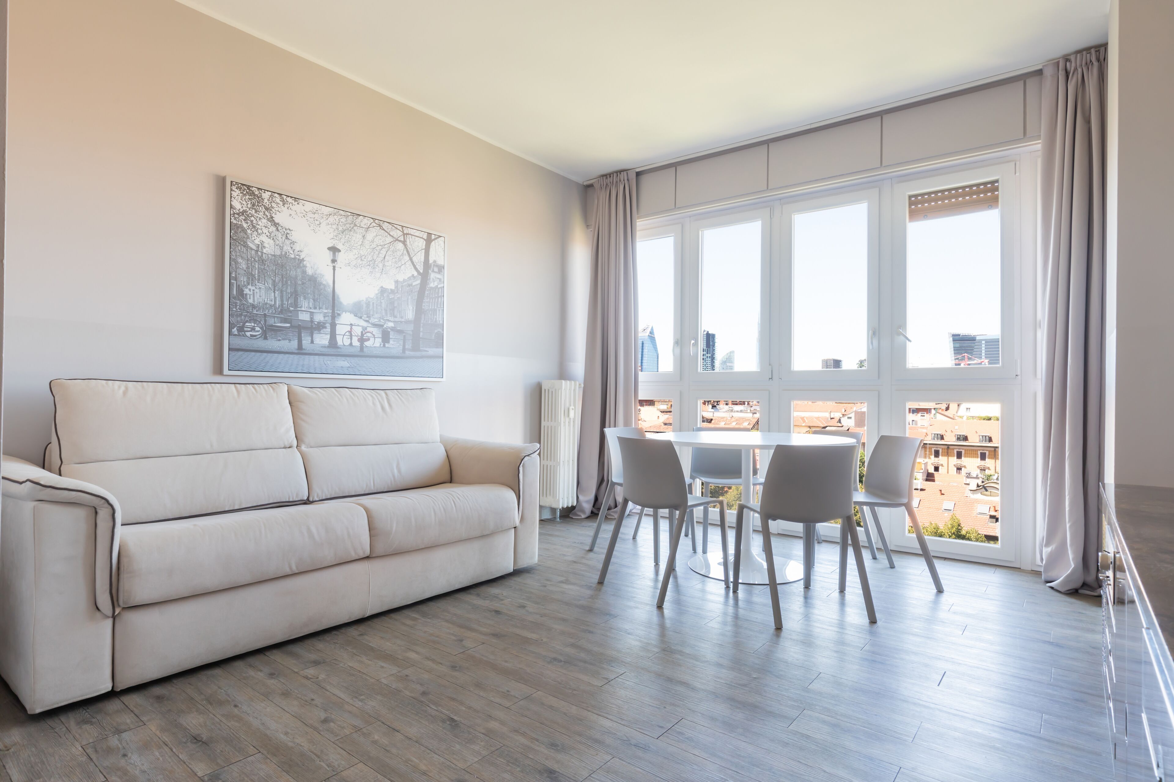 Una sala de estar moderna con un sofá color beige, una mesa de comedor con sillas, un ventanal con cortinas y vistas a la ciudad.
