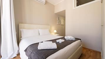 Uma cama bem arrumada com lençóis brancos e um cobertor cinza, um abajur e um ar condicionado fixado na parede.