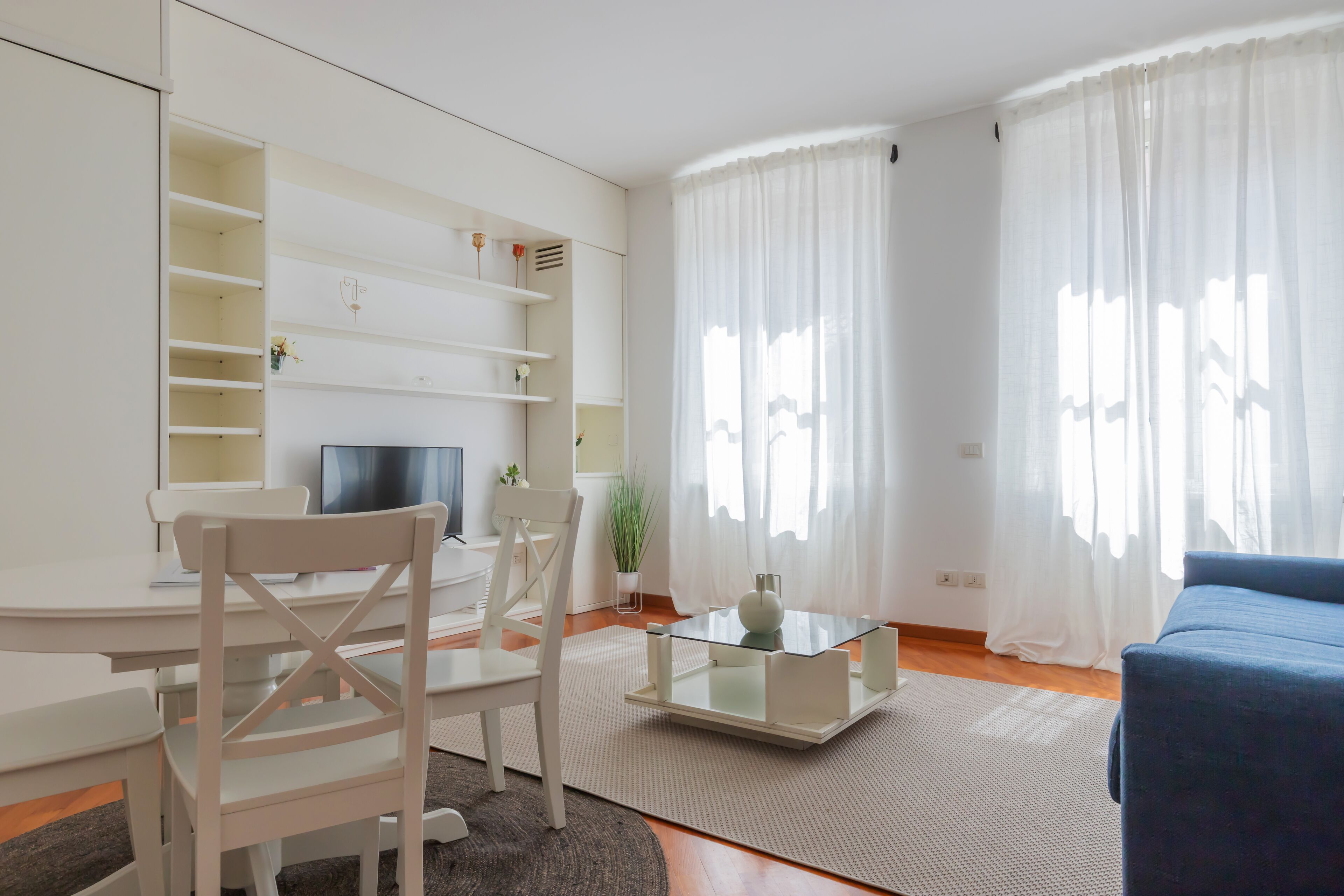 Un salon moderne avec un canapé blanc, une table basse en verre et un téléviseur posé sur un meuble. On trouve des étagères blanches au-dessus du canapé et une plante sur le sol.