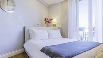 Un lit bien fait, avec du linge de lit blanc et un plaid gris, une lampe de chevet et un miroir au mur.