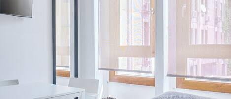 A modern room with a white table, a chair, a sofa, a TV mounted on the wall, and large windows with wooden frames.
