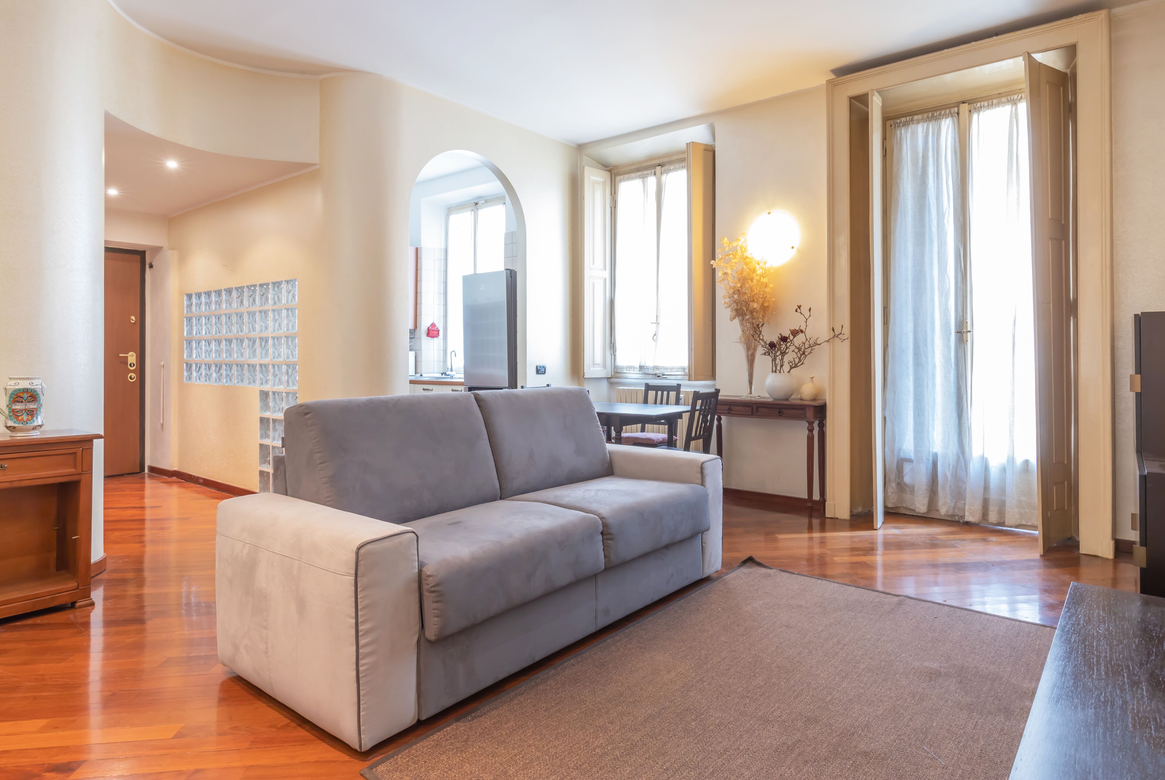 A modern living room with a grey sofa, wooden flooring, a dining area, and a kitchen visible through an archway.