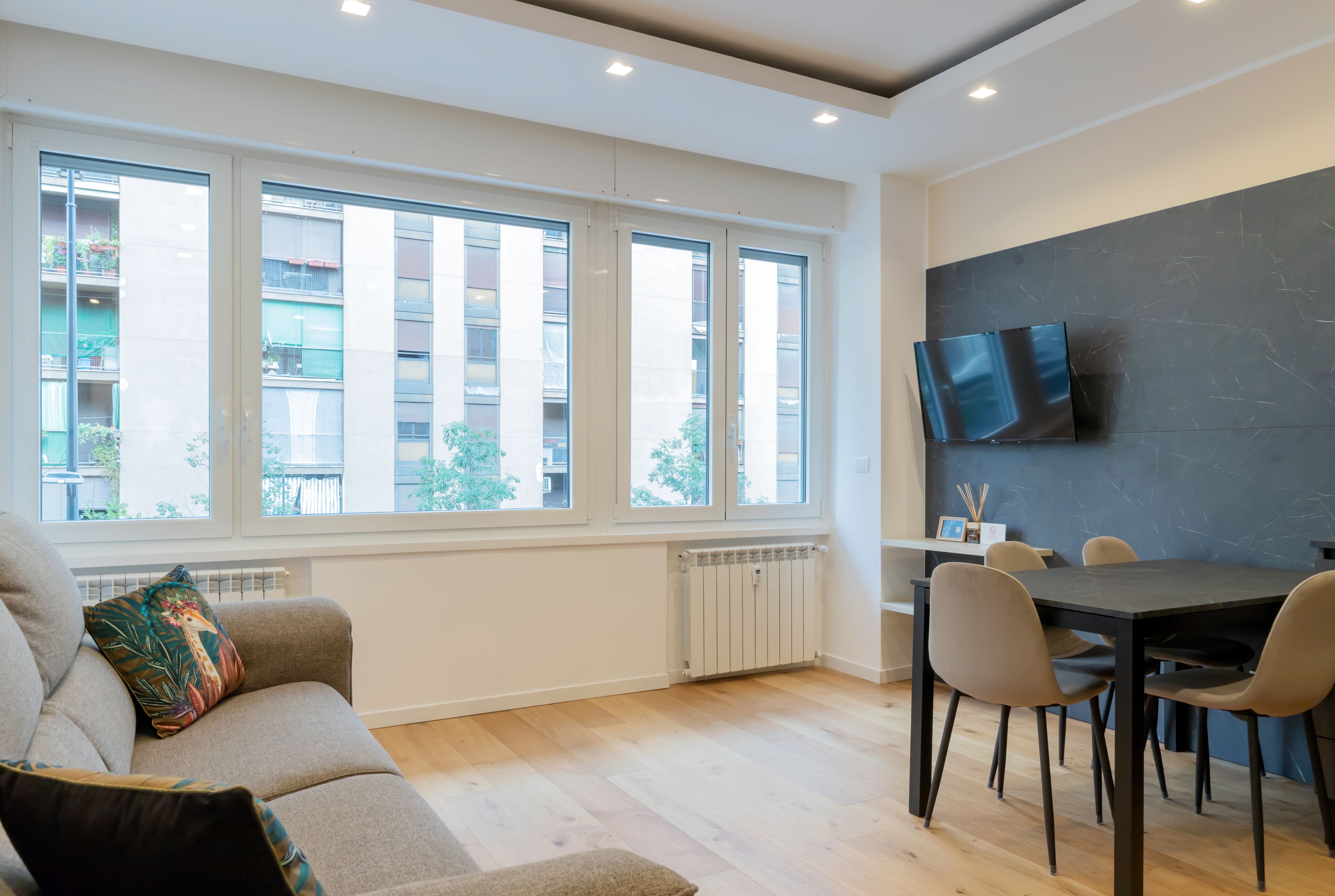 Un salon moderne comprenant un canapé, une table à manger avec des chaises, un téléviseur à écran plat fixé à un mur foncé, et de grandes fenêtres offrant une vue sur des immeubles et des arbres.