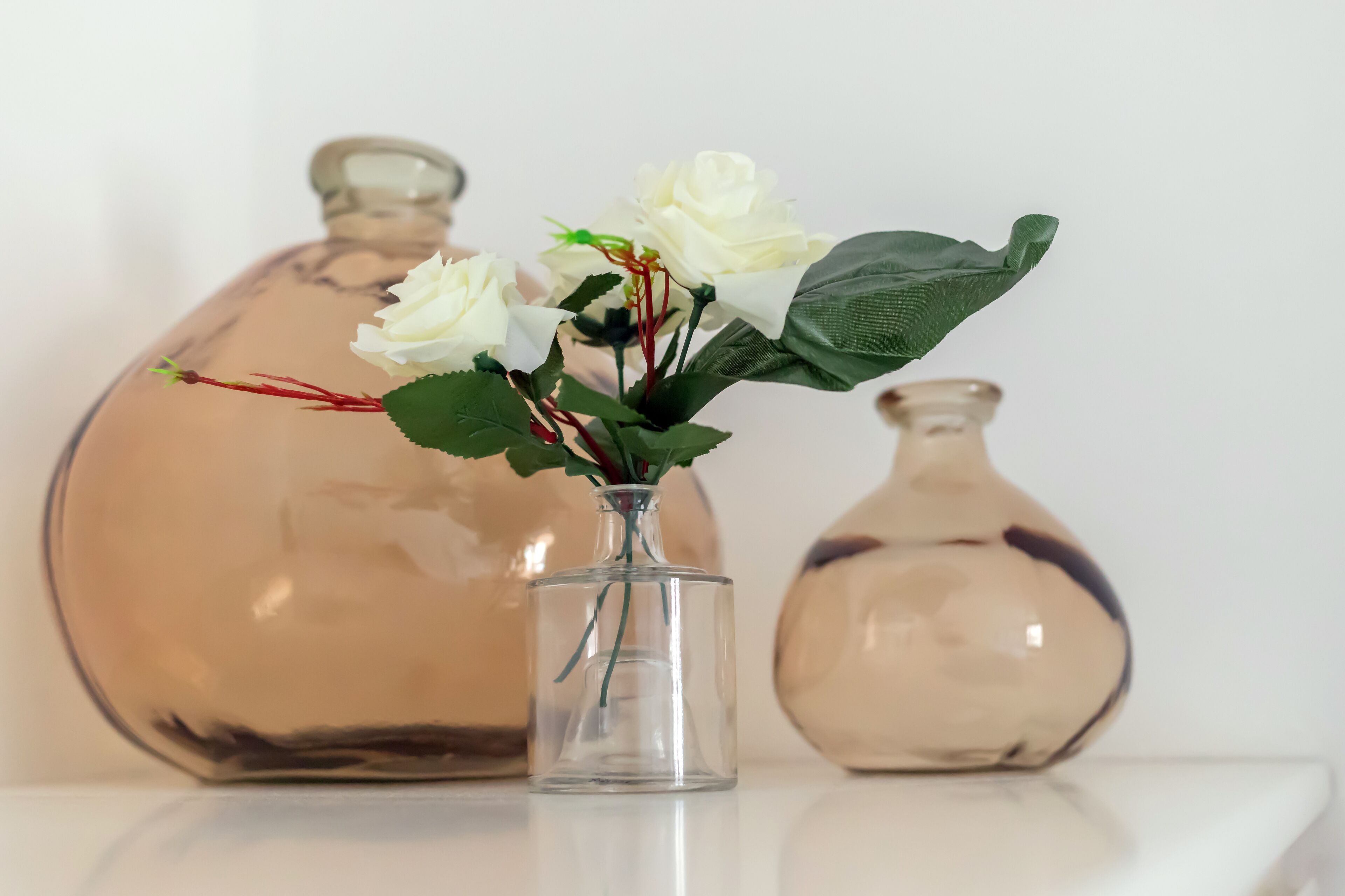 Un florero de vidrio transparente con flores blancas y hojas verdes sobre una superficie blanca.