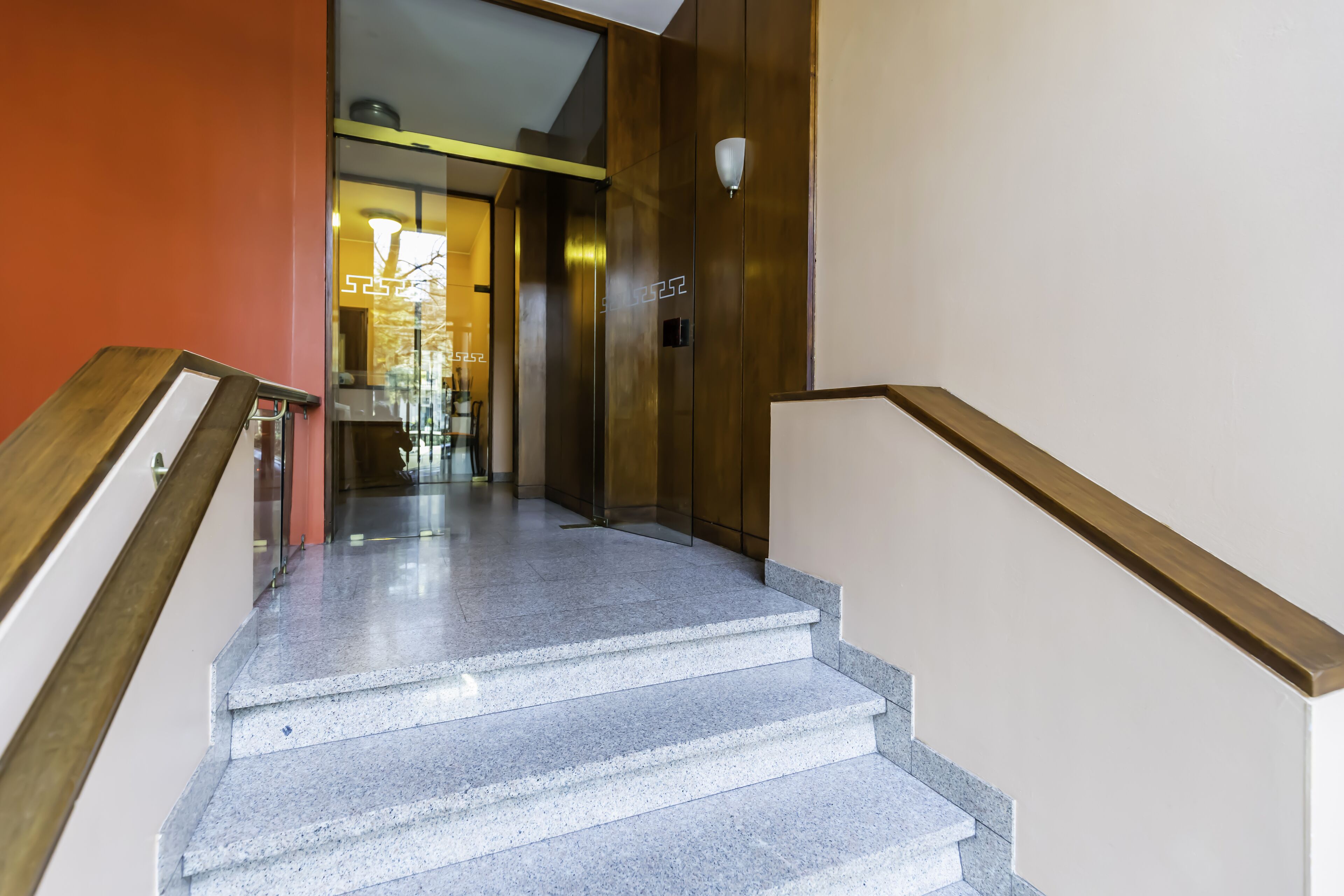 Una escalera con barandilla de madera y un portillas de vidrio en la parte superior que da a una habitación con una cama y una mesita de noche a la vista.