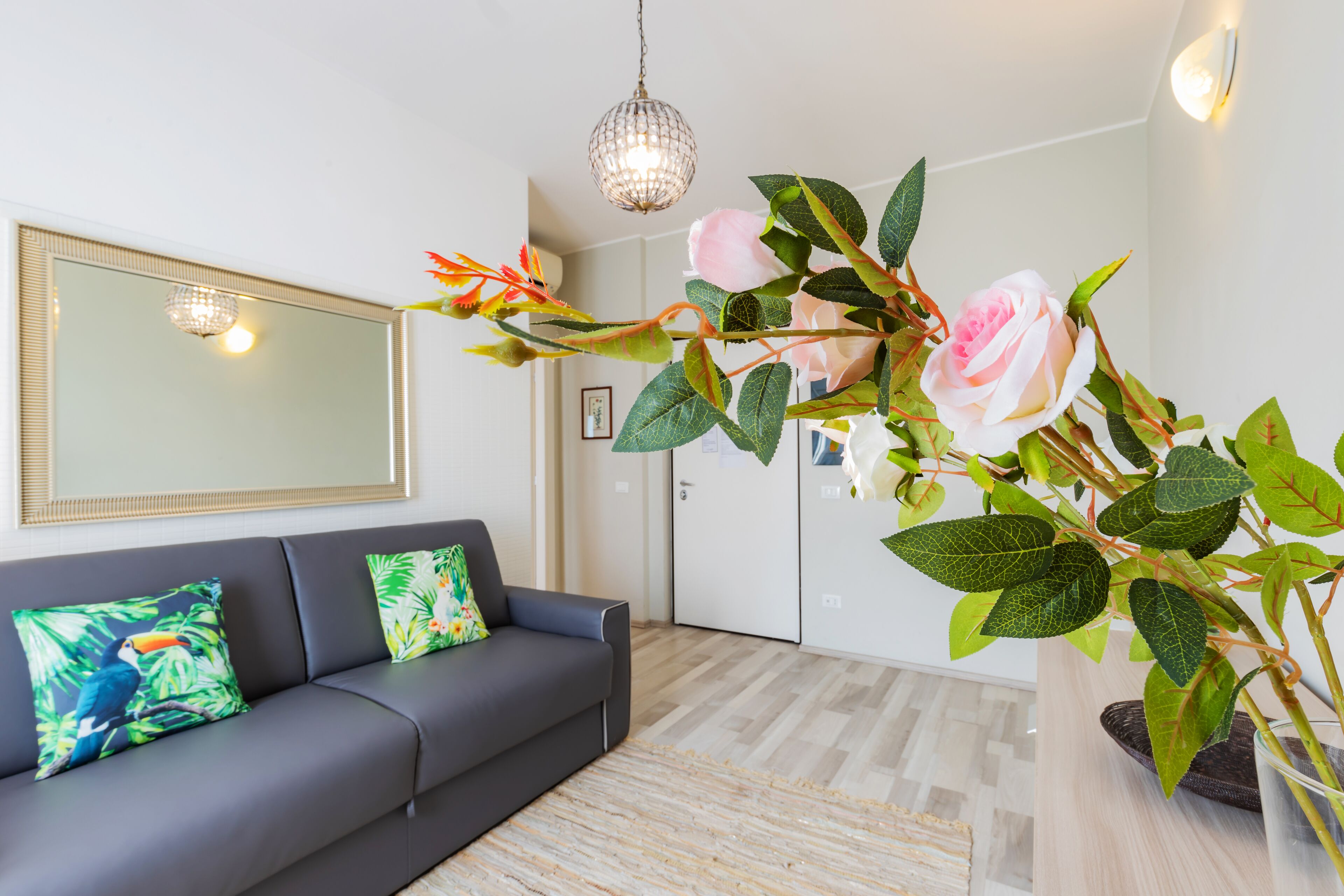 A modern living room with a grey sofa, decorative pillows, a large mirror, and a potted plant with pink flowers.