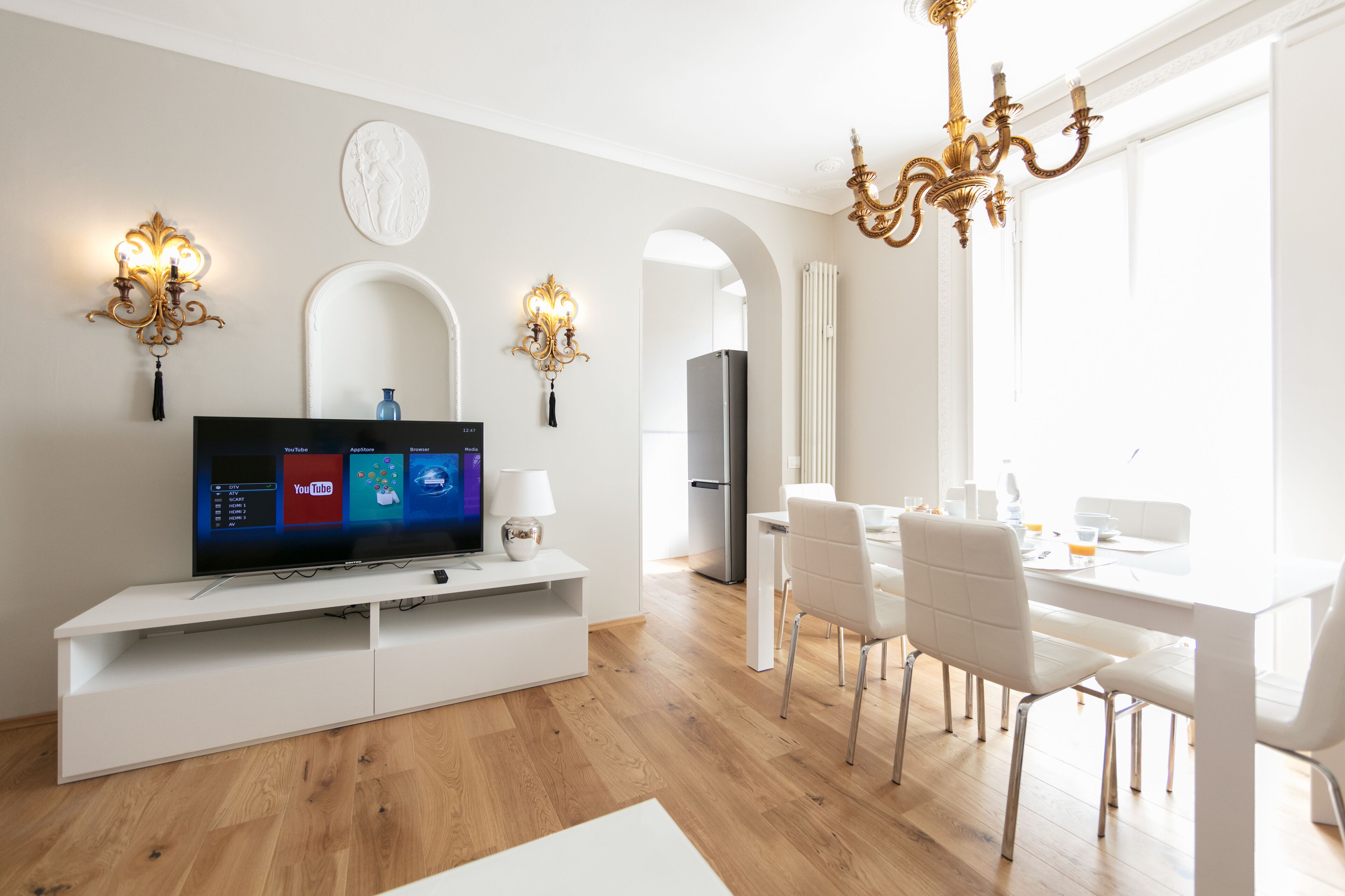 A modern living room with a flat-screen TV, white furniture, and a dining area with a chandelier.