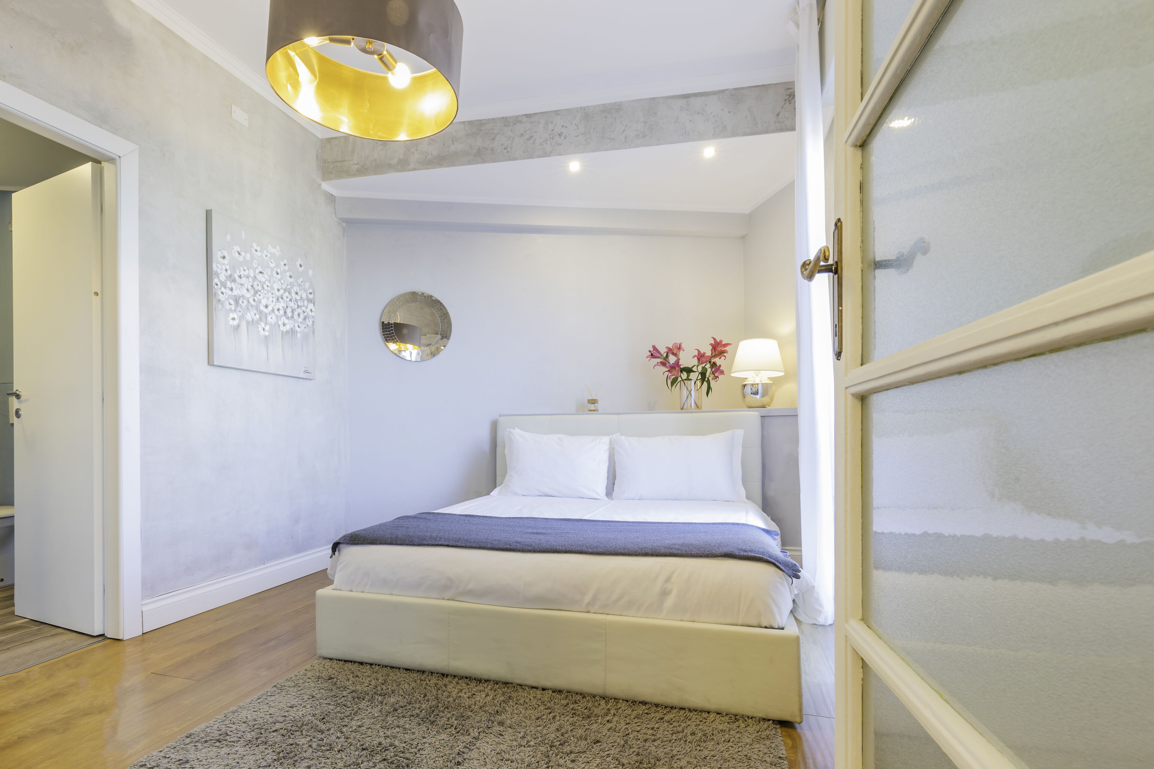 A bedroom with a bed, a bedside table with a lamp, a wall clock, and a door with frosted glass panels.