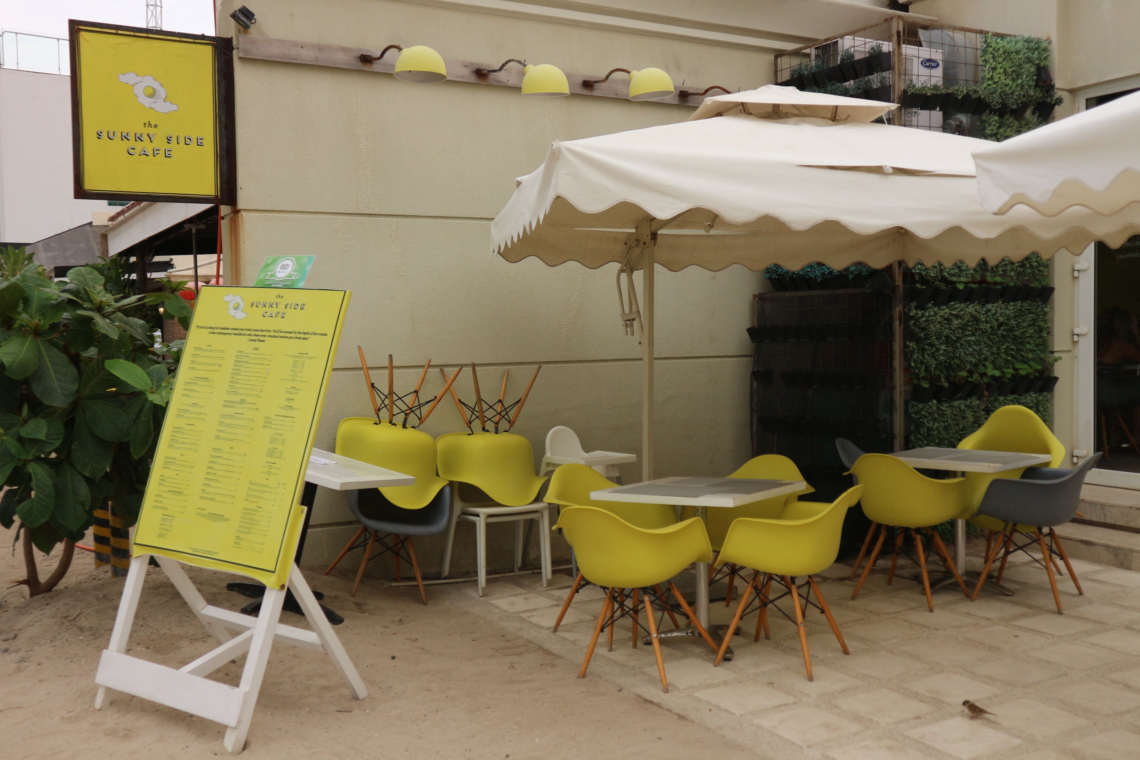 Un café al aire libre con sillas amarillas y blancas, un cartel de menú y un letrero amarillo para 'The Sunny Side Cafe'.