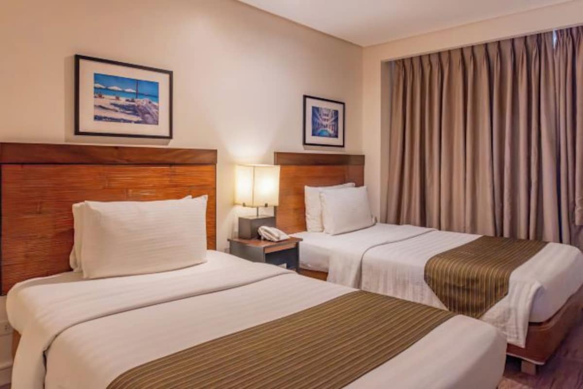 Habitación de hotel con dos camas, cabecero de madera, mesita de noche con lámpara y un ventanal con cortinas.