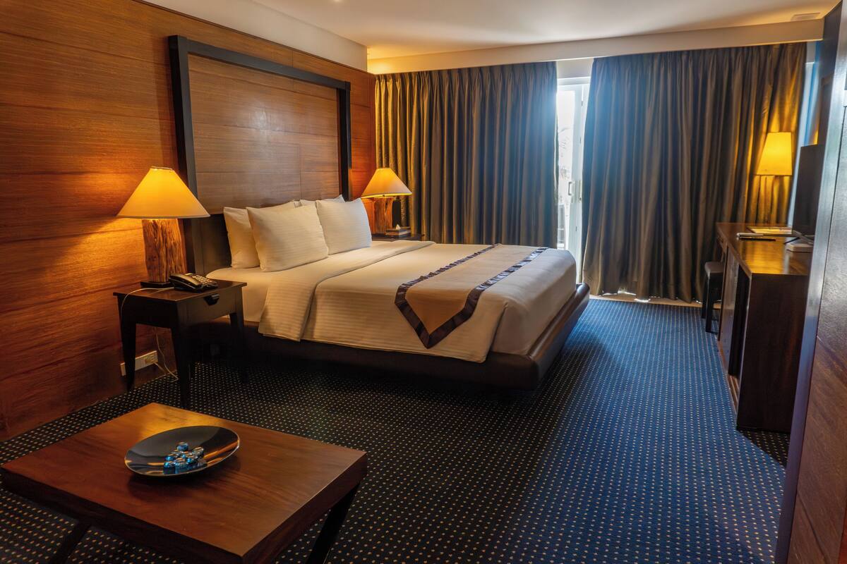 Una habitación de hotel con una cama grande, dos mesitas de noche con lámparas, un cabecero de madera, una pequeña mesa de madera con un plato decorativo y una alfombra azul con un estampado de lunares.