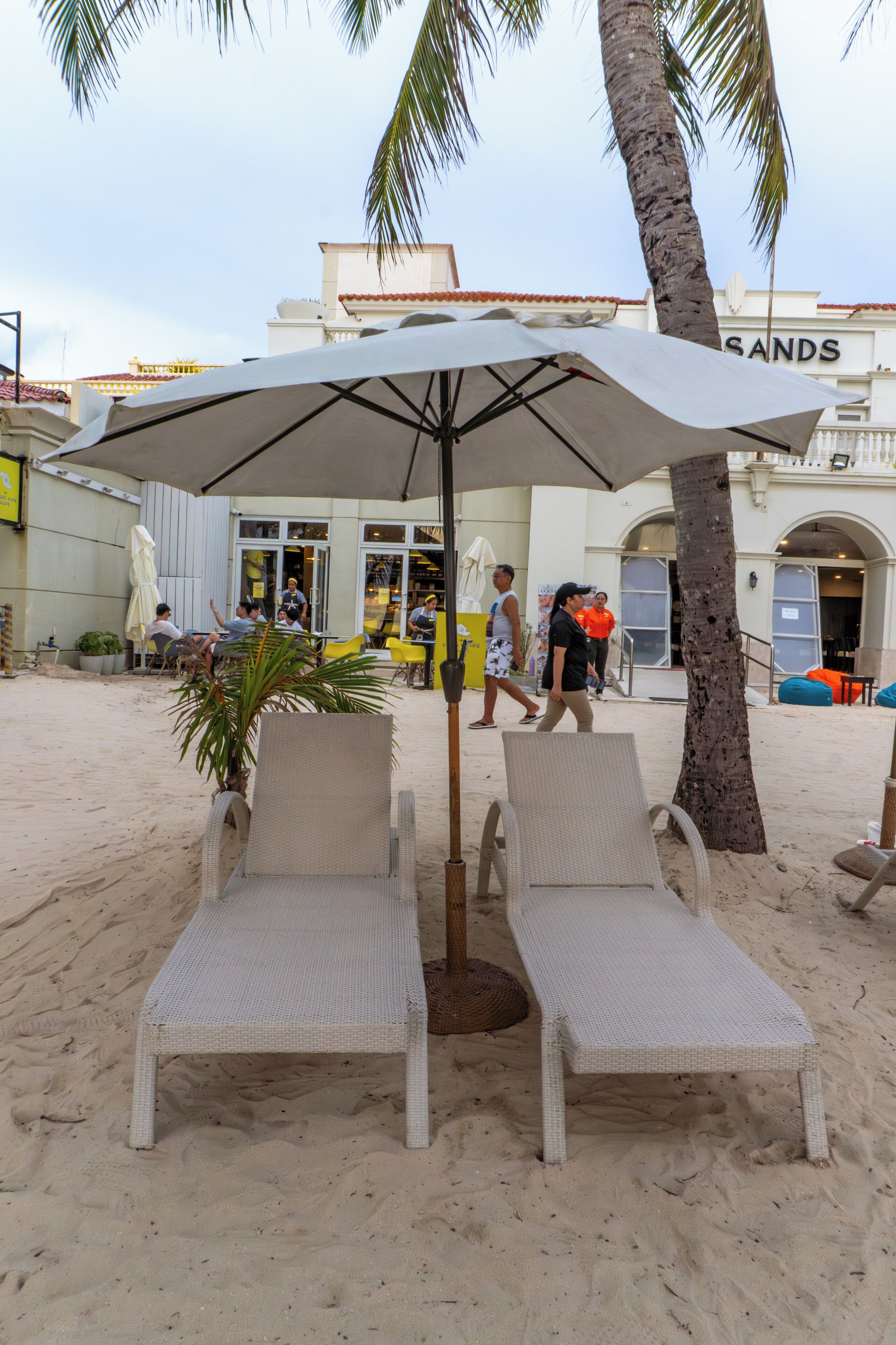 Dos sillas de playa bajo un paraguas grande, una palmera y un edificio con el letrero 'SANDS' al fondo.