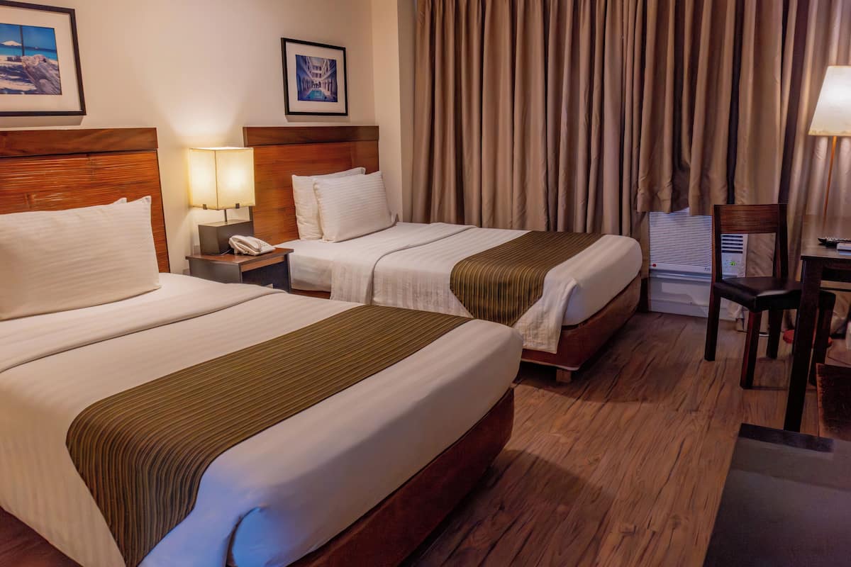 Habitación de hotel con dos camas, cabecero de madera, mesita de noche con lámpara, una silla y un escritorio con lámpara.
