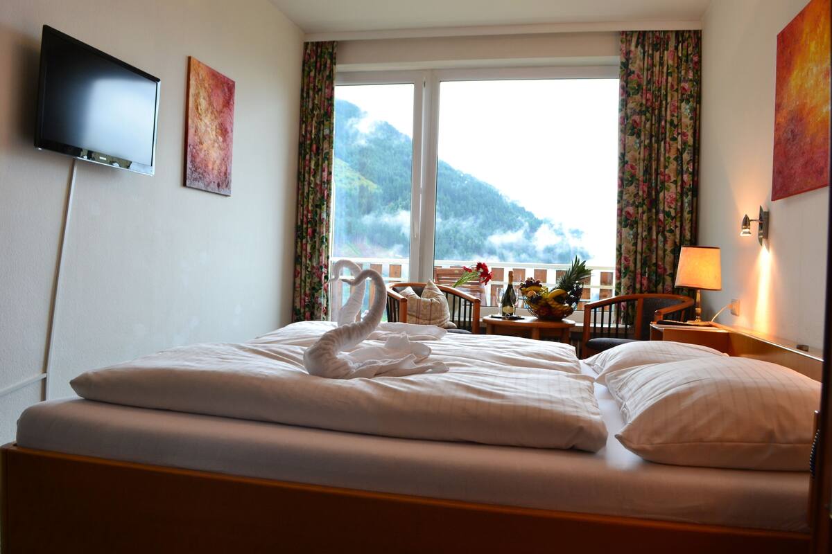 A hotel room with a bed, a TV, a window with curtains, and a view of a mountain.