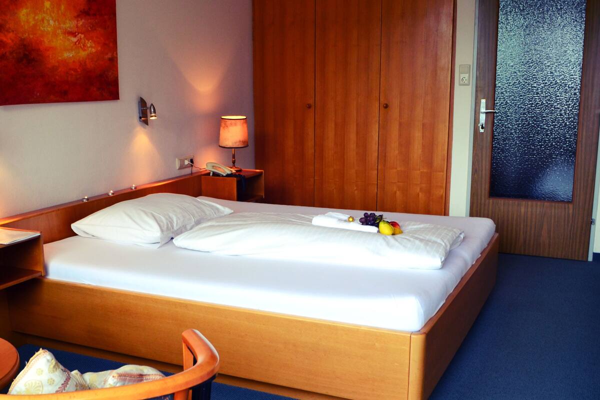 A hotel room with a bed, bedside lamps, a nightstand, a vase with flowers, a bottle of wine, and a fruit arrangement on the bed.