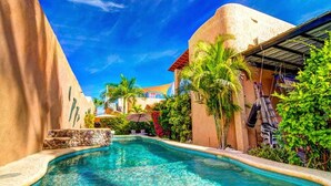 A pool area with a curved pool, palm trees, and a building with a red roof in the background. - Casa Kootenay Waterfront BnB (La Paz)