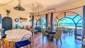 A room with a round dining table, chairs, a hanging light fixture, and a view of the sea through large windows. - Casa Kootenay Waterfront BnB (La Paz)