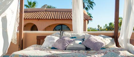 A bed with patterned bedding, a wooden headboard, and a view of a building with a tiled roof and arches.