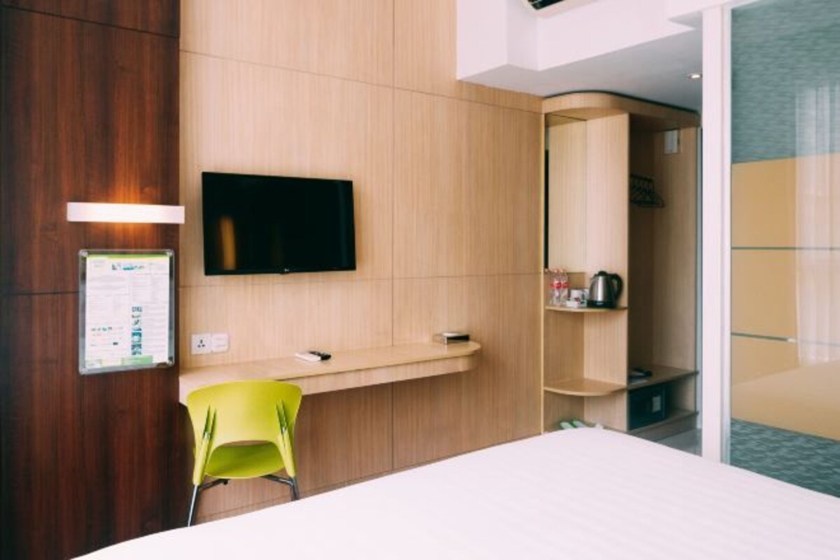 Une chambre d’hôtel avec un mur en bois, un téléviseur à écran plat fixé au-dessus d’un bureau, une chaise jaune et une petite étagère contenant des articles de toilette.