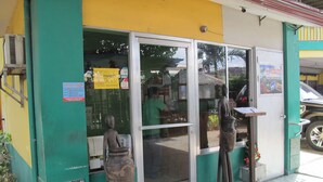 Un edificio con fachada verde y amarilla, entrada por puerta de vidrio y un pequeño espacio exterior con una estatua.