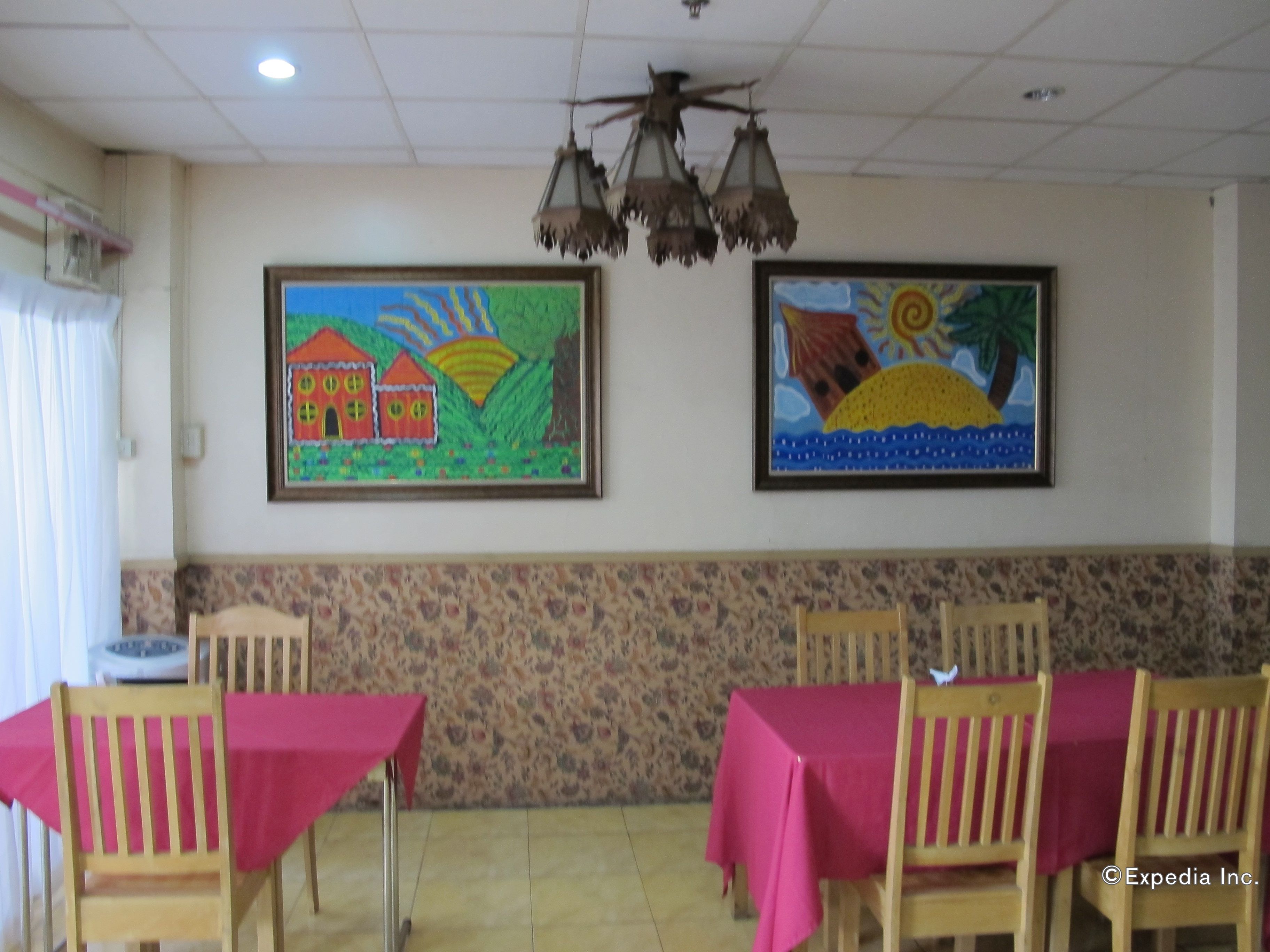 A dining area with wooden chairs and tables covered with red tablecloths, two framed paintings on the wall, and a ceiling fan.