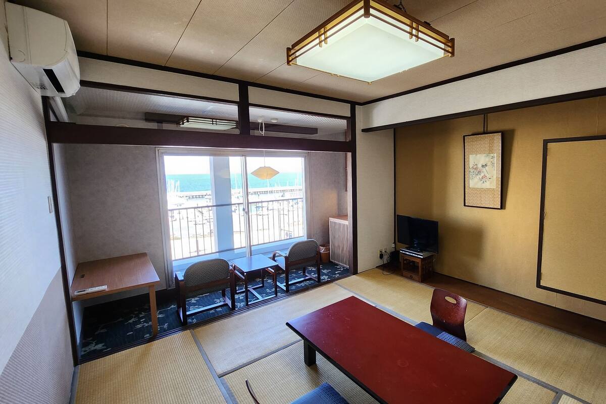 Una habitación japonesa tradicional con suelo de tatami, una mesa baja, sillas, un televisor y vista al mar a través de amplios ventanales.