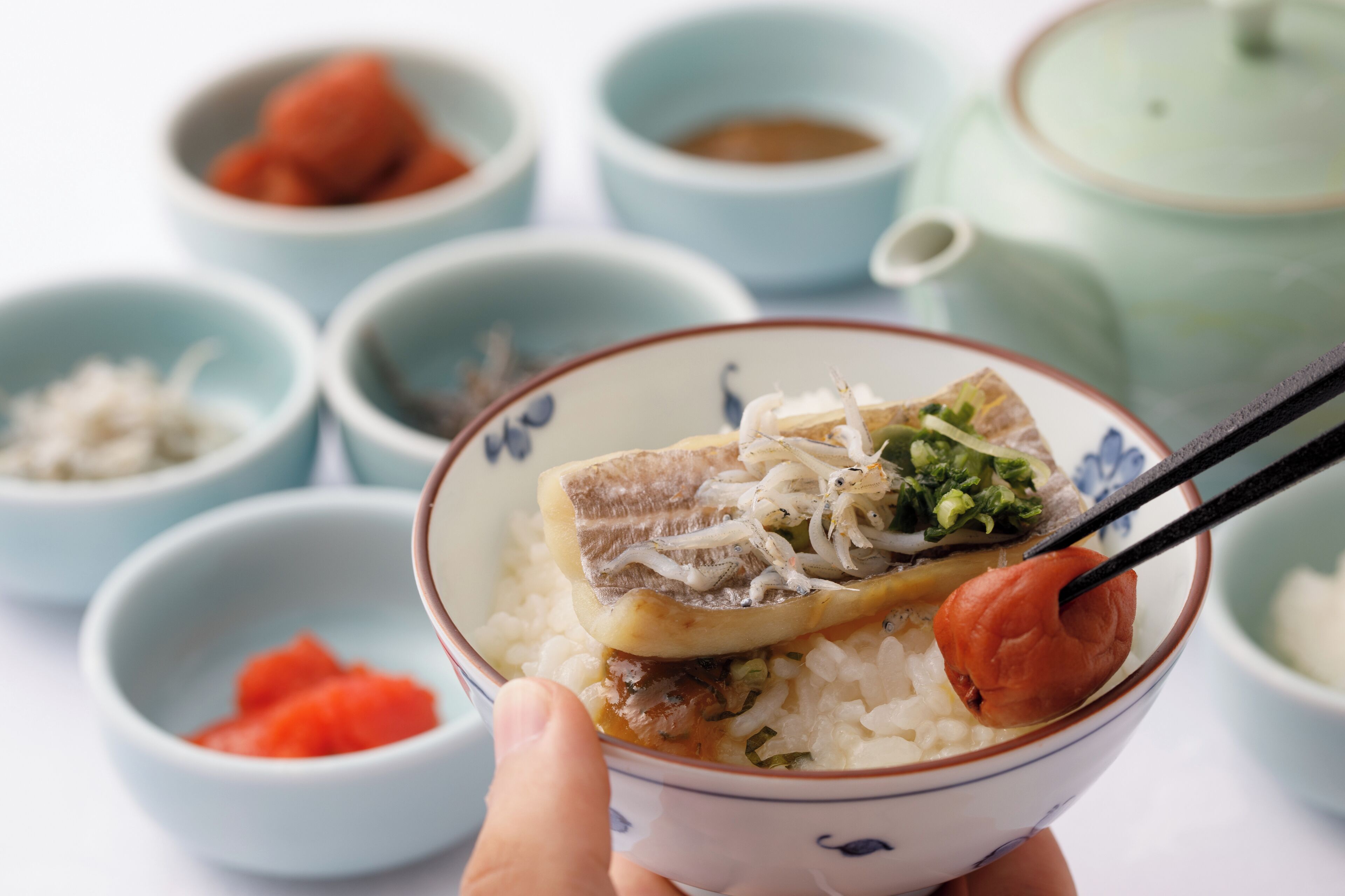 A bowl of rice with fish, vegetables, and a piece of meat, accompanied by small bowls of condiments and a teapot.